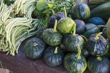 Vibrant outdoor market stall displays fresh harvest of green kabocha squash, long bean, and other healthy vegetable. This raw agricultural abundance evokes natural feeling