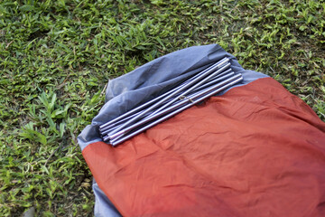 Folded orange camping tent with an aluminum pole on green grass background. This outdoor equipment shows preparation and anticipation for hiking adventure or travel