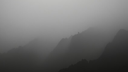 The silhouetted peaks of majestic mountains, enveloped in a thick, atmospheric, and ethereal blanket of dense gray mist, create a mesmerizing monochrome landscape during the early morning or twilight.