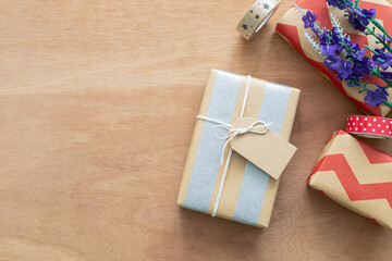 Top view of beautifully wrapped gift boxes with kraft paper and brwon tag for text on a light wooden surface. A bundle of purple flowers and decorative tapes add a festive and creative touch.