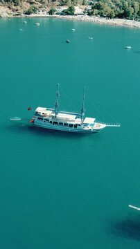 Vertical drone video capturing a serene white two-masted yacht with a Turkish flag floating in turquoise waters near Kekova Bay. Ideal for mobile and social media storytelling.