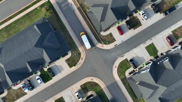 Residential America suburb with a yellow school bus turning through a clean four-way junction surrounded by modern homes, driveways and neatly arranged lots in daylight. Aerial top down tracking.