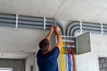 Electrician installing corrugated pipes and connecting colorful electrical wires to a fuse box mounted on a concrete wall and ceiling during building construction and renovation