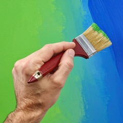Close-up of a human hand holding a paintbrush, applying paint to a surface that is part green and part blue