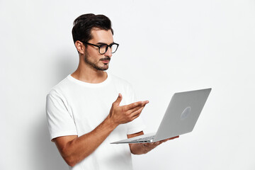 Thoughtful man wearing glasses and white t-shirt stands holding a laptop while gesturing. Isolated on plain solid light background. Modern technology concept.