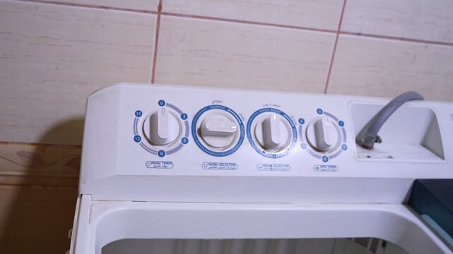 Close-up of a man's hand adjusting the selector dial on a top-loading washing machine control panel. Features labels in both English and Arabic for wash settings.