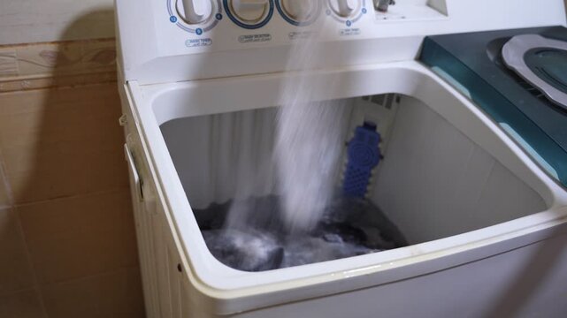 Close-up of white granular laundry detergent powder being poured into a top-loading washing machine filled with clothes. Authentic domestic laundry routine.