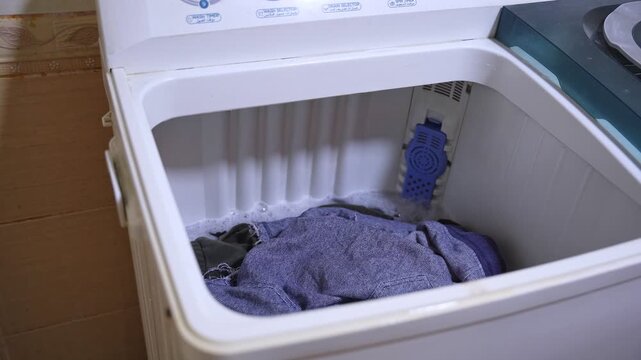 Close up view of a hand reaching towards dark clothes spinning in a white top-loading washing machine. Authentic domestic laundry routine with soapy water and motion.