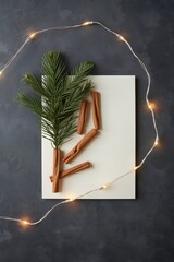Festive holiday composition with blank paper, evergreen branch, cinnamon sticks, and warm string lights on dark textured background for seasonal greeting or invitation design
