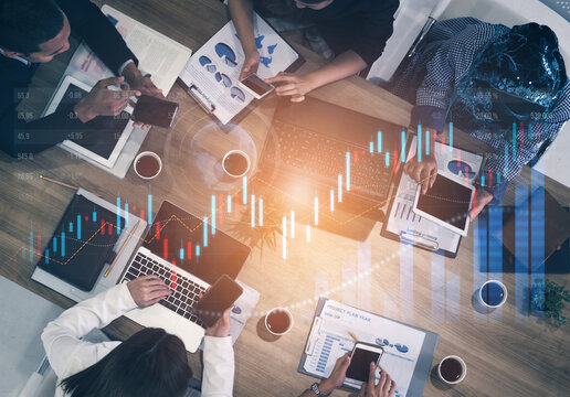 A dynamic business meeting setting showcases professionals analyzing data and discussing stock market trends with laptops and mobile devices present. Scalp