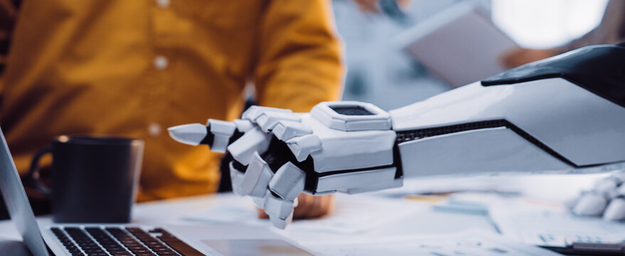 A robotic arm reaches towards a laptop in a busy office setting, symbolizing the intersection of technology and teamwork in modern work environments.Noogenesis