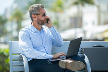 Confident business man sitting on a bench with a laptop and phone in park. Successful business man. Mature businessman enjoying work break. Businessman taking a break on a park bench. Summer business.