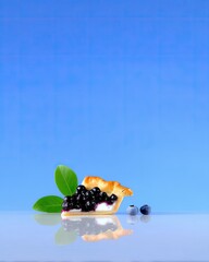 A slice of blueberry pie with fresh blueberries and green leaves, reflected on a shiny surface, against a bright blue background.
