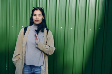 Woman in beige coat and gray sweater stands before a green metal wall, casual outdoor street...