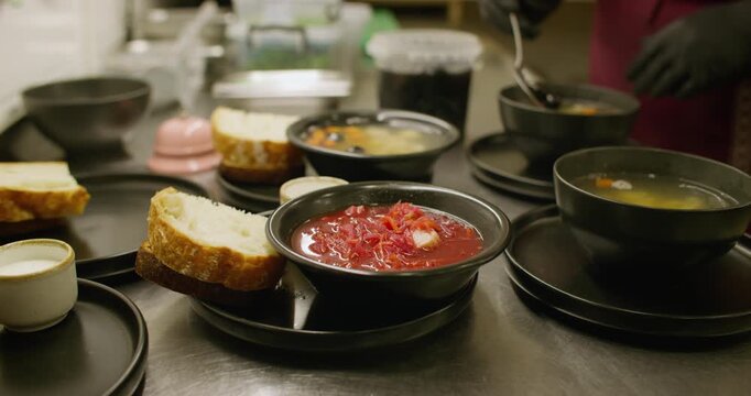 Catering cooking. Cafeteria lunch. Chef adding olives to hot steaming soup on kitchen table. Serving borsch in black bowl with bread.