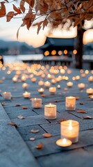 Close-up of numerous lit candles arranged on a stone surface, with autumn leaves and a blurred background of lights and a structure, evoking a sense of tranquil