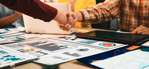 A diverse business team celebrates a successful partnership with a handshake over various financial reports and data analytics in a bright modern office environment. SACTR