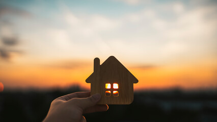 A hand holding a wooden house model against a warm sunset sky symbolizes home, dream living, hope, and real estate concepts. Soft golden light enhances themes of security, family, and future lifestyle
