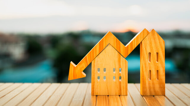 Wooden house and building models with a downward arrow symbolizing falling real estate prices, market decline, and financial downturn. Soft sunset light enhances themes of risk, investment challenges,