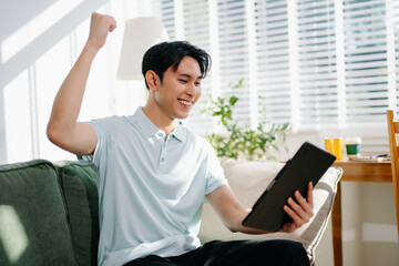 Young asian man relax on comfortable couch at home texting messaging on smartphone and tablet smiling man use cellphone shopping online from home