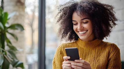 Woman with curly hair smiles while looking at her phone in front of a bright window with a plant