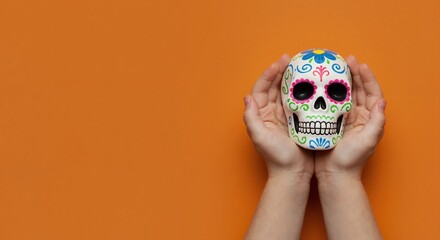Hands holding traditional Mexican sugar skull for Dia de los Muertos. Colorful calavera decoration on orange background with copy space