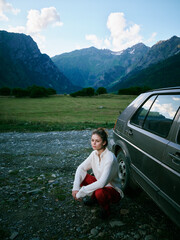 Obraz premium Woman crouching beside a car in a mountain valley, lush meadow, distant peaks, blue sky, tranquil outdoor travel scene, adventure and exploration in a scenic alpine landscape