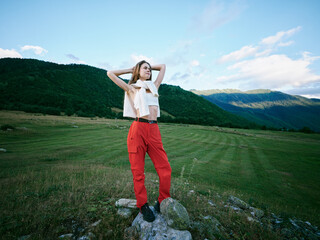 Obraz premium Outdoor field with mountains in the distance, a fashion pose on a rocky ledge, bright sunlight, red pants and white top, conveying freedom, adventure and a carefree summer mood in wide open space