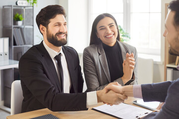 Happy smiling couple shaking hands reaching agreement with a man realtor or financial advisor signing successful contract sitting at desk on workplace. Insurance agent consulting clients in office.