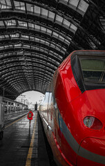 Solo traveler in red coat walking beside sleek high speed train under Milan central station roof rainy travel photography cinematic perspective leading lines wanderlust and Europe journey