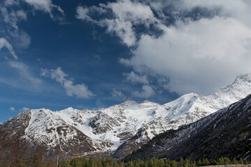 Obraz premium Wide scenic view of snowy highlands with towering peaks, shifting clouds and sunlit slopes, capturing calm, scale and raw beauty of untouched alpine nature in winter light