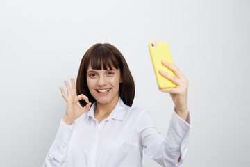 Cheerful woman in a white shirt takes a self portrait with a bright yellow smartphone, making the ok gesture while posing against a clean neutral background.