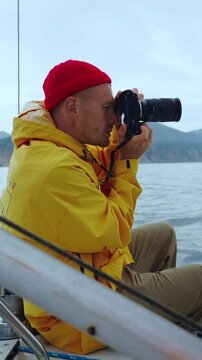 Capturing Moments: A Dedicated Photographer in a Bright Yellow Rain Jacket and Red Beanie Focuses on His Camera While Sailing in Scenic Waters