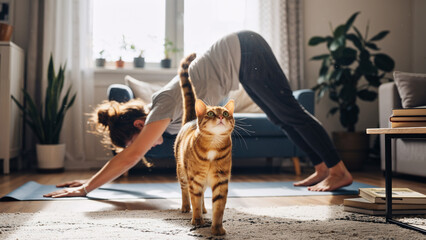 Woman doing yoga on mat at home with an interested ginger cat. Pet friendly fitness and healthy lifestyle. Relaxation exercise.