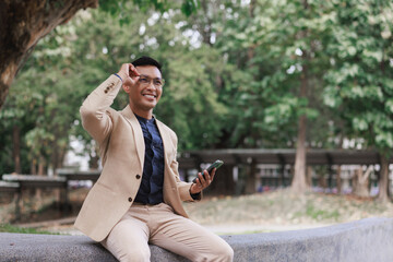 Smiling asian man adjusting glasses while using smartphone in park