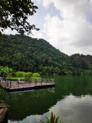 Fototapeta premium A wooden floating dock extending into a calm green lake backed by a dense, forested hill. The scene is tranquil and quiet. Use it for lifestyle websites, travel campaigns, or environmental reports.