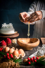 Hands cracking an egg into a wooden bowl with Christmas baking ingredients, festive cupcakes, and holiday decorations. Warm seasonal cooking atmosphere perfect for holiday food themes.