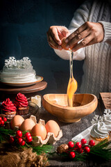 Hands cracking an egg into a wooden bowl with Christmas baking ingredients, festive cupcakes, and holiday decorations. Warm seasonal cooking atmosphere perfect for holiday food themes.