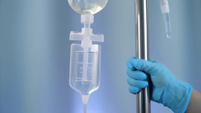 Close-up of a medical professional's gloved hand holding an IV pole with a saline drip bag.