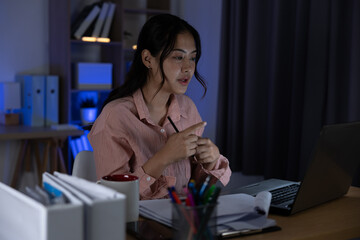 Asian young woman working at night in home office speaking on laptop while holding pen and notes