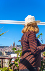 Obraz premium Woman with her back to the camera, wearing a straw hat, on a sunny rooftop overlooking the city of Barcelona, ​​with the Sagrada Familia in the background.