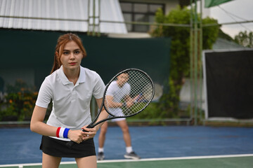 A young White tennis player stands in a ready position at the net, holding her racket firmly while a teammate prepares behind her