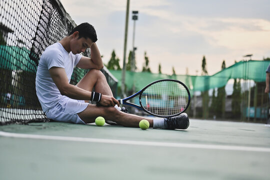 A tired tennis player relaxes on the court at dusk, sitting with his racket and cooling down after a demanding practice