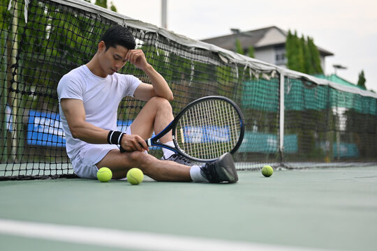An Asian male tennis player sits beside the net after a difficult practice moment, reflecting on performance and regaining focus