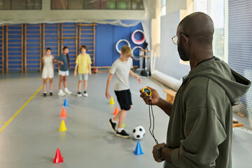 Black man coach timing group of Caucasian boys and girls teenagers dribbling soccer ball through cones during sports training in gymnasium, focusing on athletic skill development