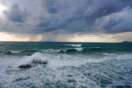 Dramatic view of strong wind and high waves crashing over rocks under a dark, brooding storm sky. Intense marine landscape with a visible rain squall on the horizon.