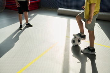 Two boys playing soccer indoors, one Caucasian child standing with foot on soccer ball while another boy facing him, both wearing shorts and sneakers on sports court