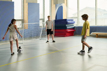 Three teenagers, two boys and one girl, playing soccer in indoor gym, Caucasian and possibly multiethnic group, girl dribbling ball while boys preparing to defend goal, casual athletic activity