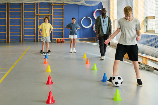 Caucasian teenage boy dribbling soccer ball through colorful cones in gymnasium while Black man coach and multiethnic teenagers observing training session in background - Powered by Adobe
