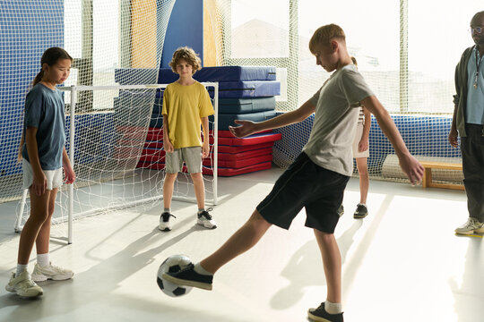 Group of multiethnic teenagers playing indoor soccer, Caucasian boy kicking ball while Asian girl and another Caucasian boy watching, adult Black man standing nearby observing action